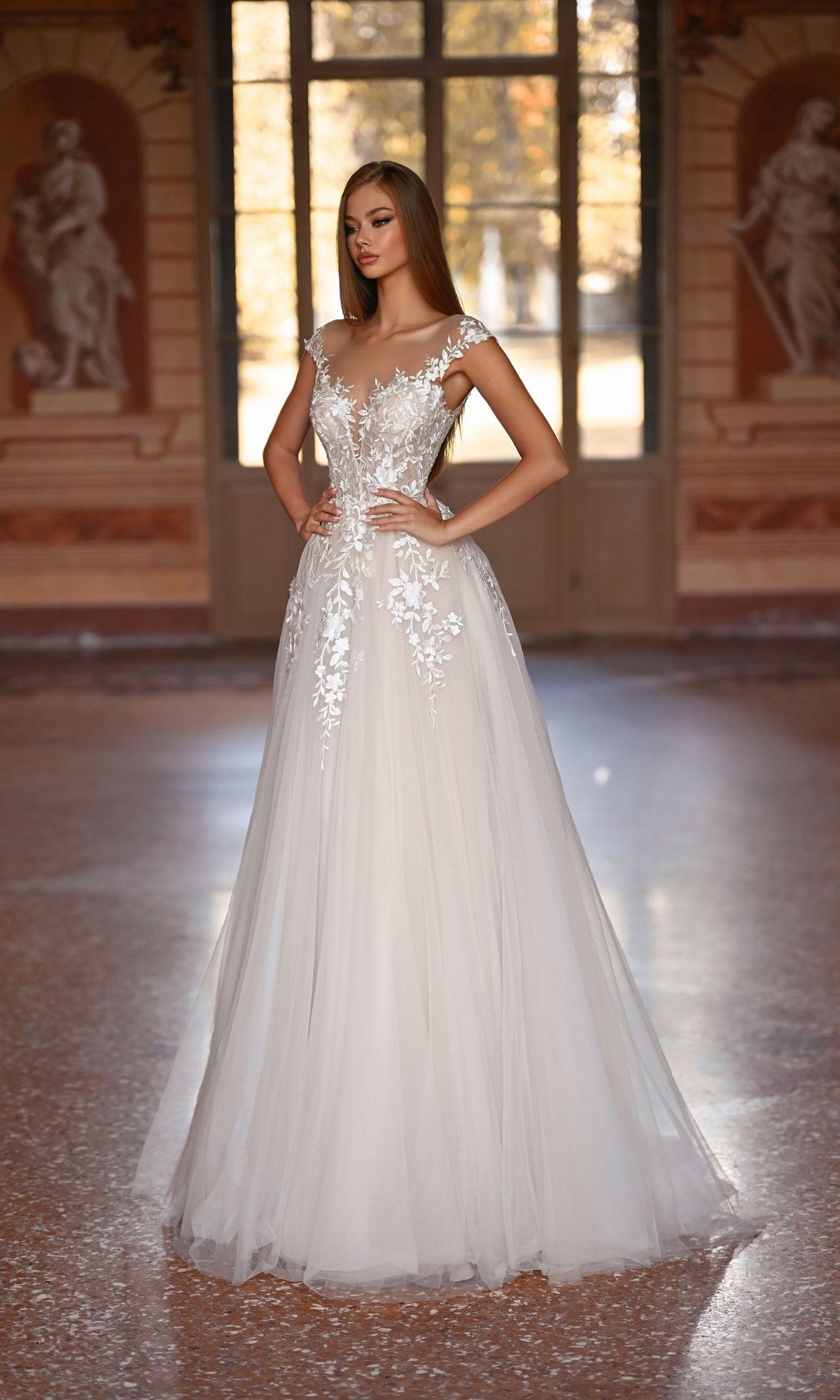 A woman wearing the Alyce 5090 long formal dress, featuring lace embroidery and an a-line tulle skirt, stands in a sunlit room with classical statues and large windows.